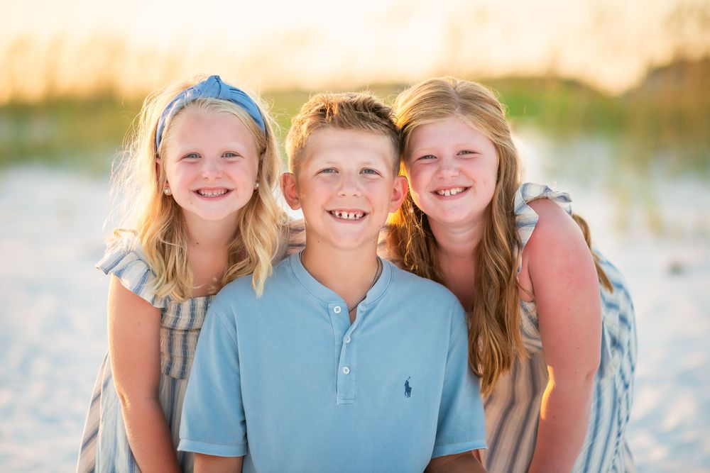 Three siblings wearing coordinated blue and white outfits pose together on a sunny beach at sunset.