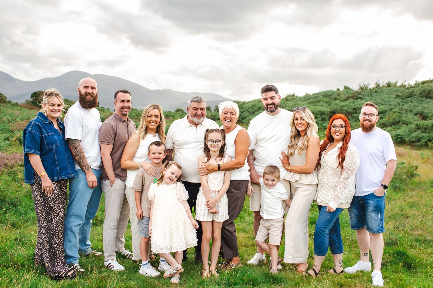 A large group in coordinated cream and white outfits poses together in a green mountain landscape.