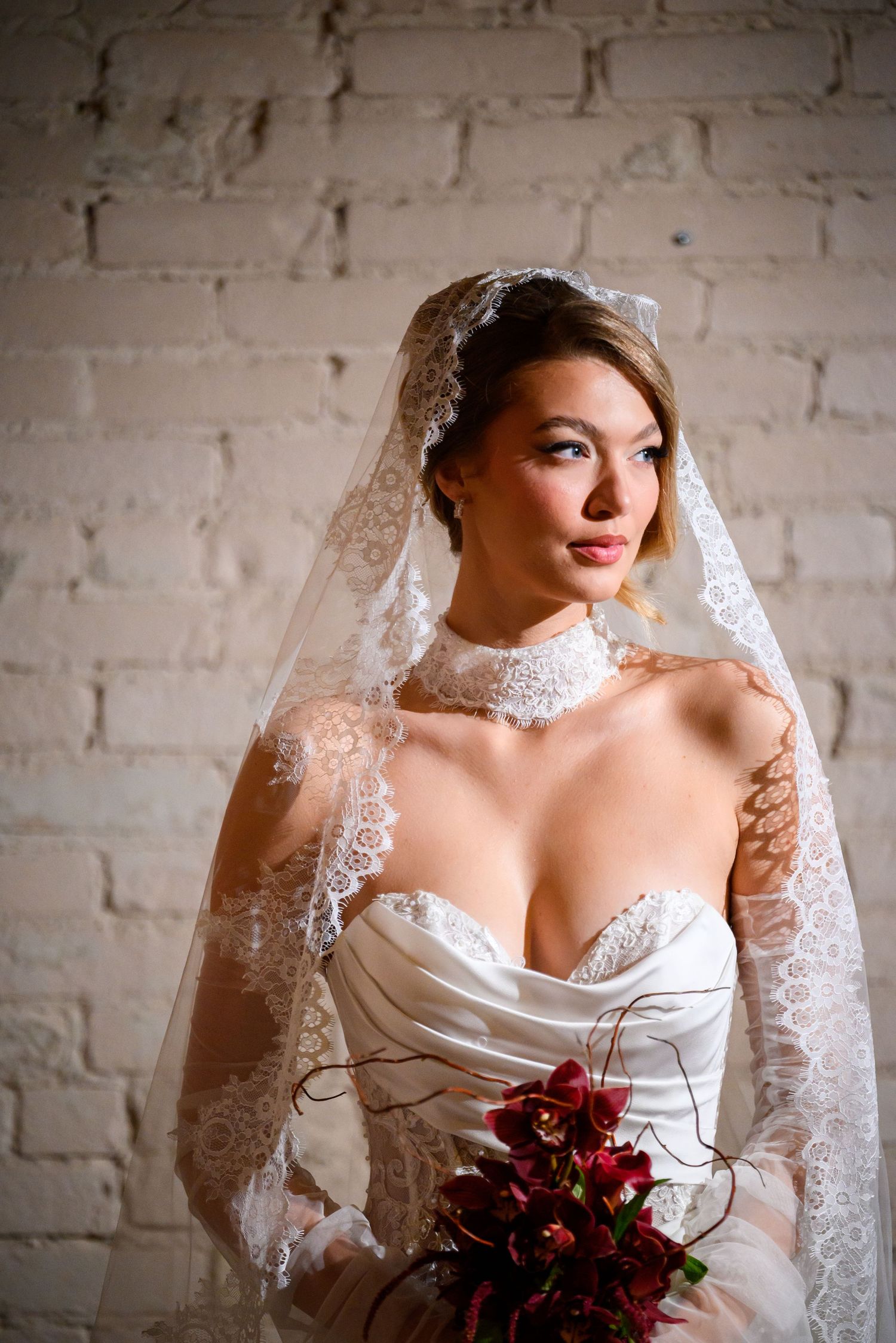 A bride in a white wedding dress with lace veil poses against a white brick wall holding a deep red bouquet.