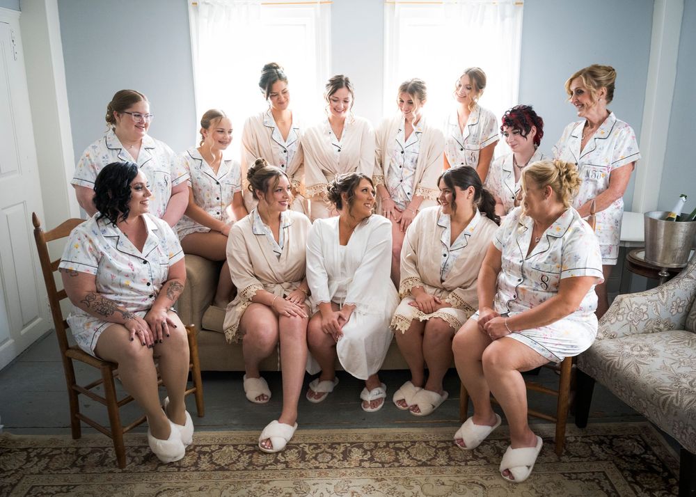 Bride surrounded by bridesmaids in matching pajamas laughing together during fun wedding morning preparations.