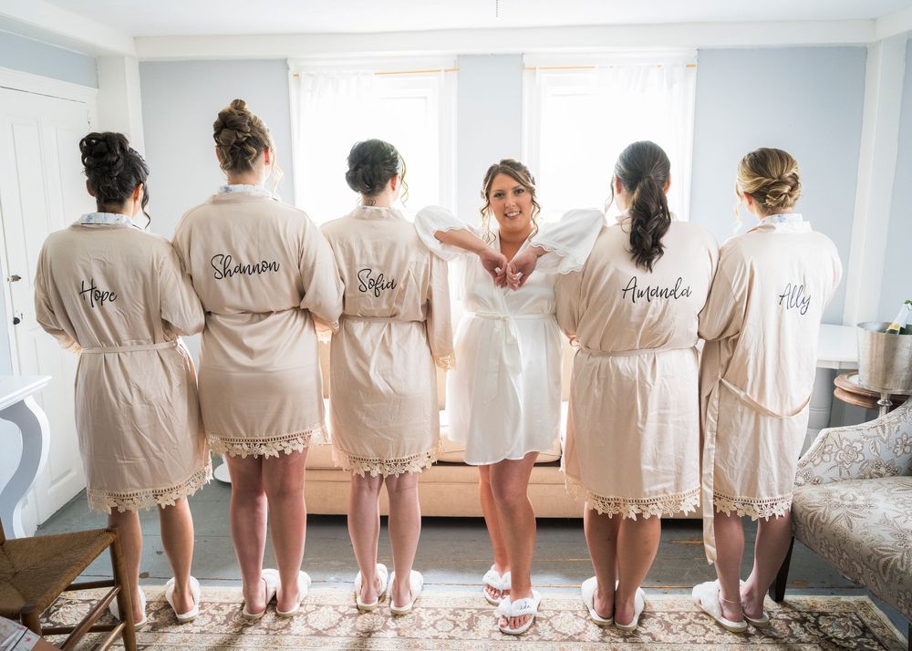 Bride poses with bridesmaids in personalized lace-trimmed robes showing embroidered names during wedding preparations.