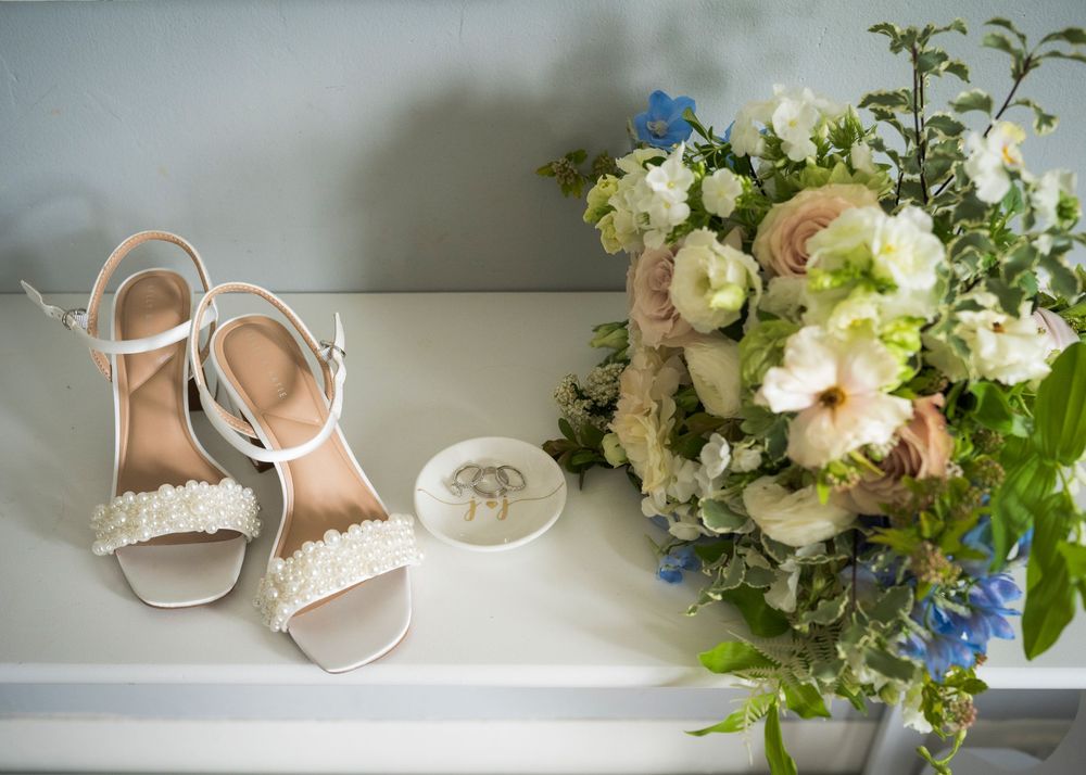 White bridal heels with pearl details beside wedding bouquet of white and blue flowers on dresser.