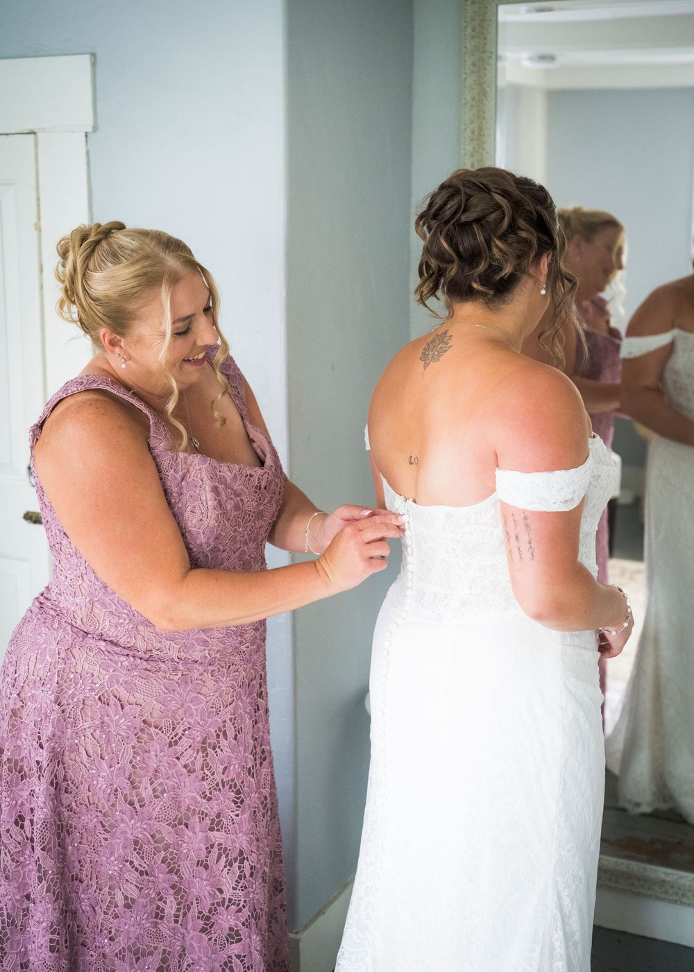 Mother in lavender lace dress helps bride button her off-shoulder white wedding gown before the ceremony.