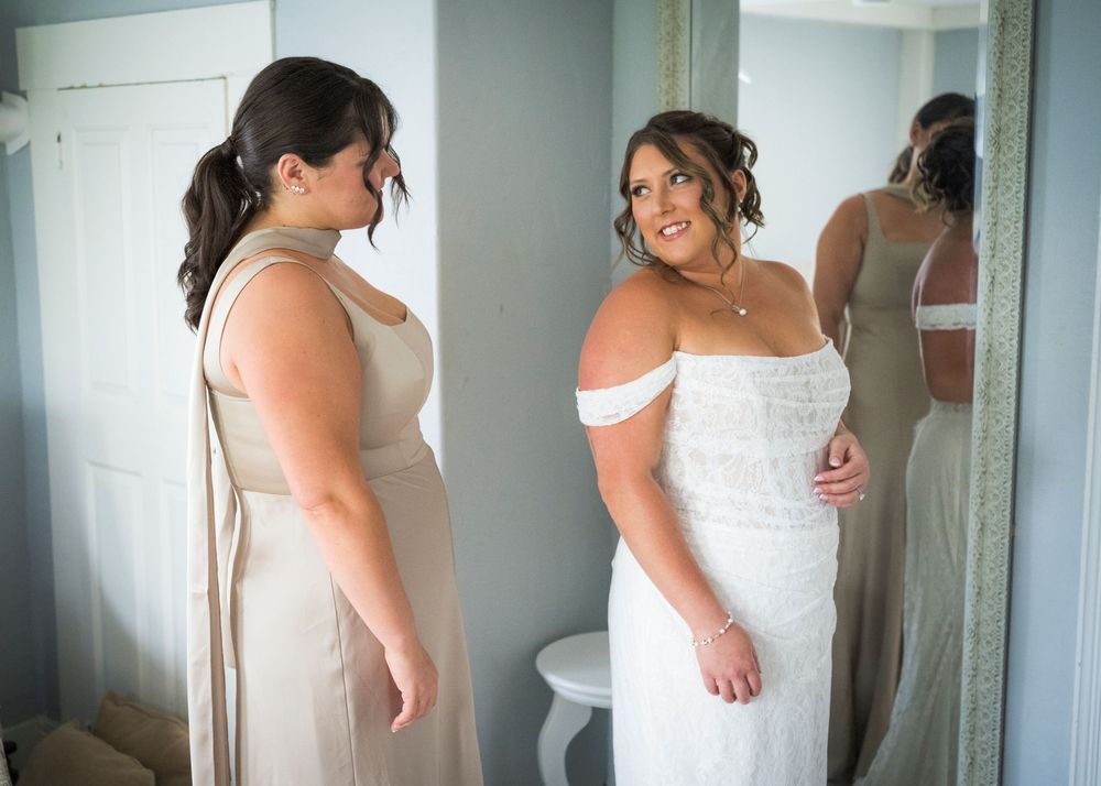 Bride in off-shoulder lace gown admires reflection in mirror with bridesmaid in champagne dress.