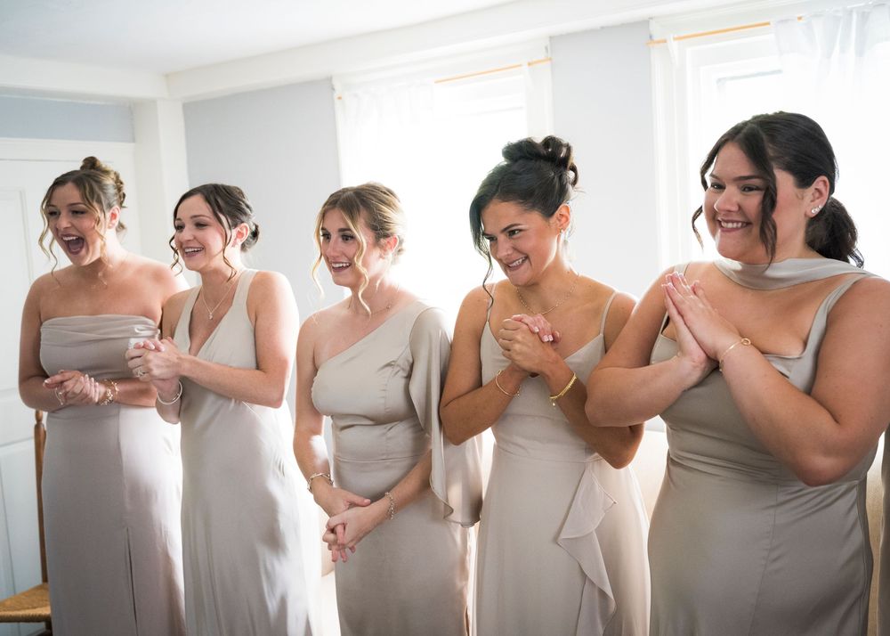 Five bridesmaids in champagne gowns react with joy while seeing the bride before the wedding.