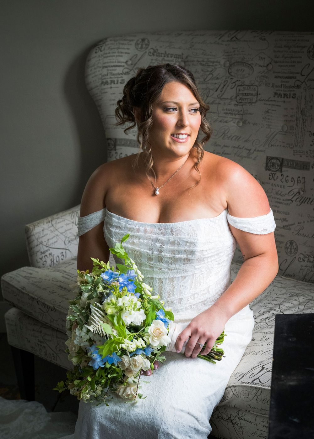 Bride in off-shoulder white gown holding blue and white bouquet seated on French script chair.