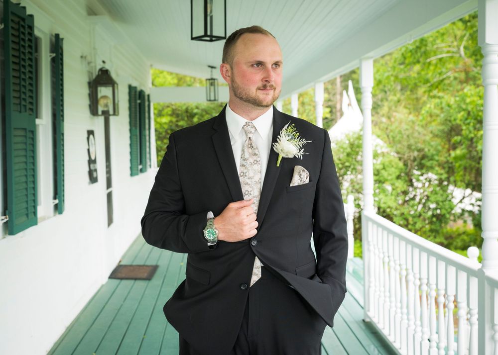 Groom in black suit with boutonniere standing on white porch before wedding ceremony.