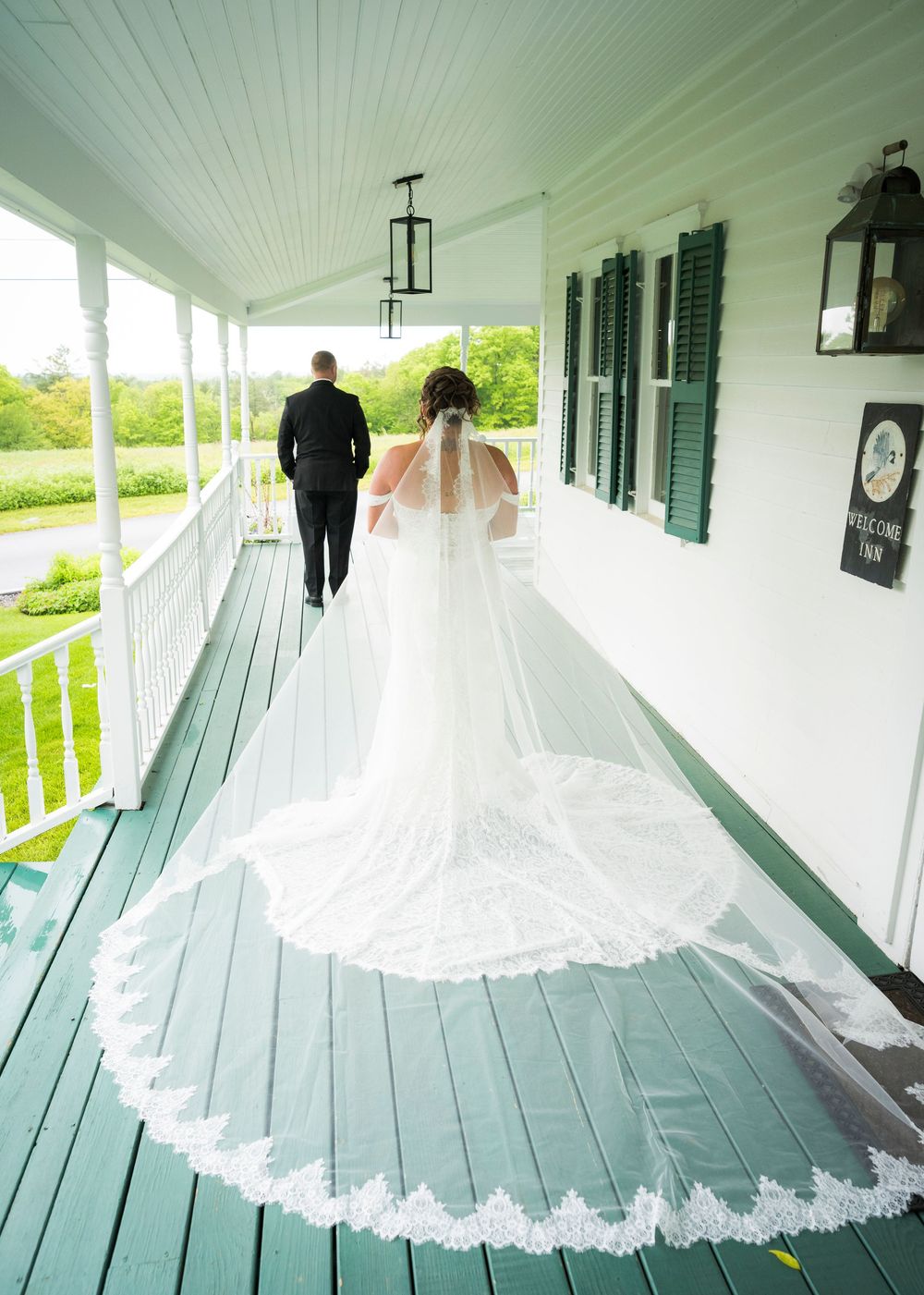 Bride in lace-trimmed cathedral veil stands with groom on white porch during first look moment.