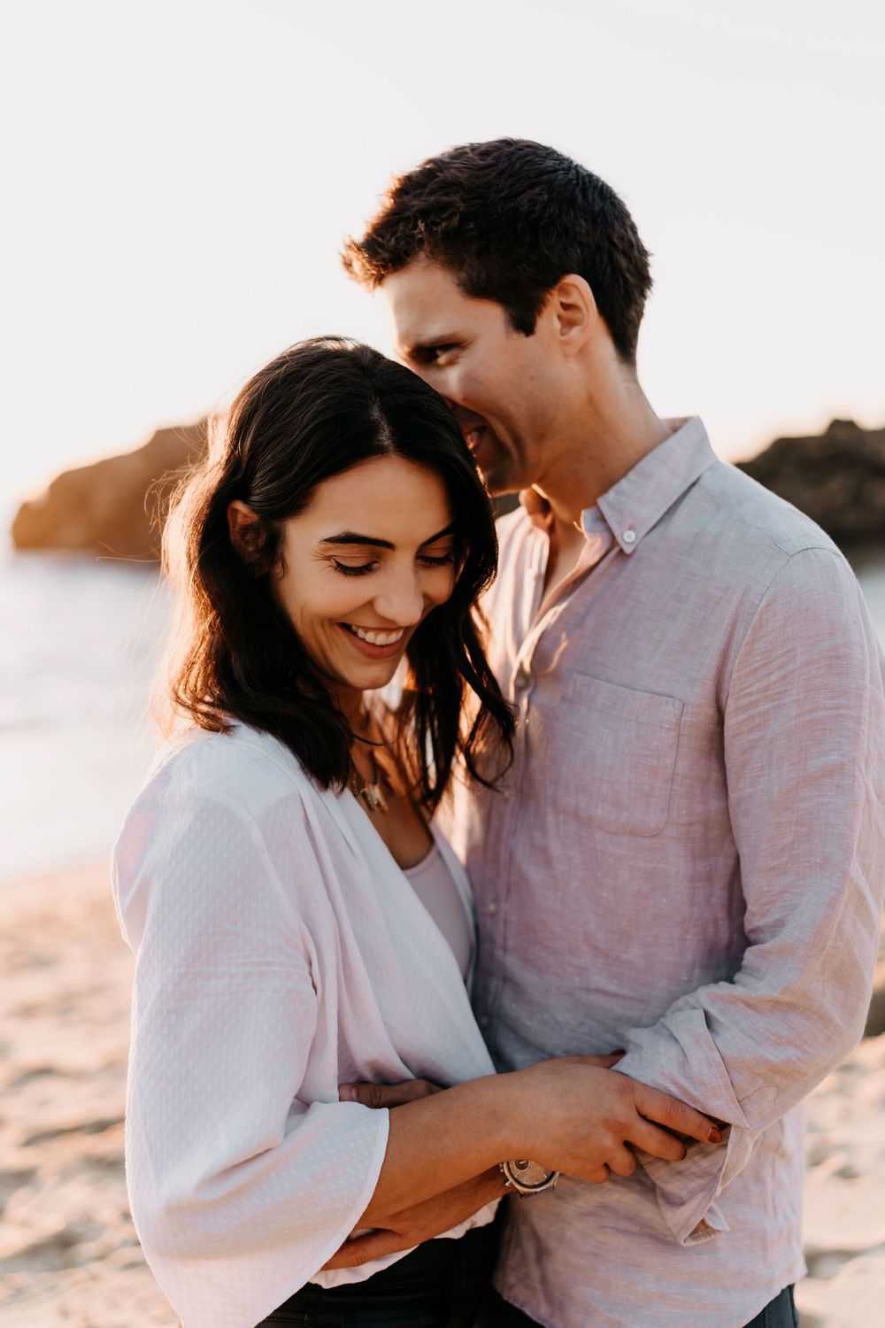 A couple embraces and laughs together during a romantic sunset photoshoot on the beach.