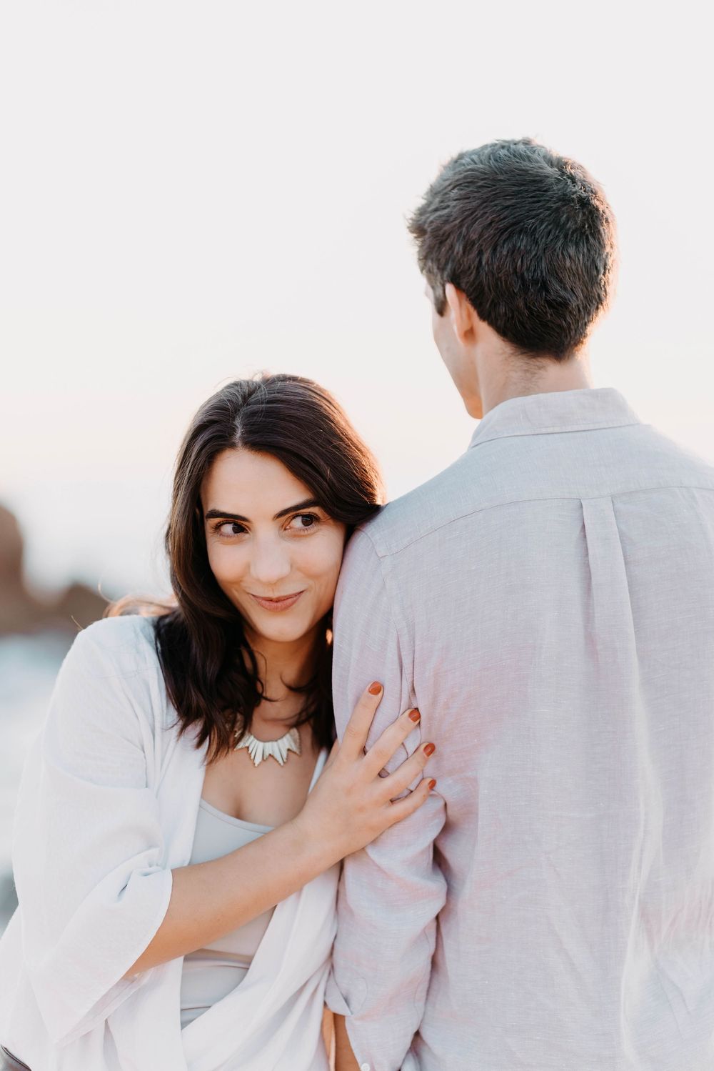 A couple embraces during a romantic sunset photoshoot, with the woman wearing white and peeking around her partner's shoulder.