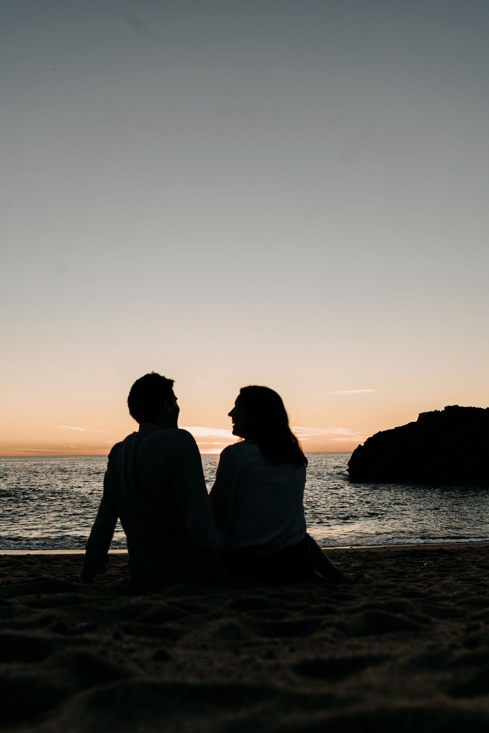 Silhouettes of two people sitting on a beach at sunset with ocean waves and rock formations in the background.