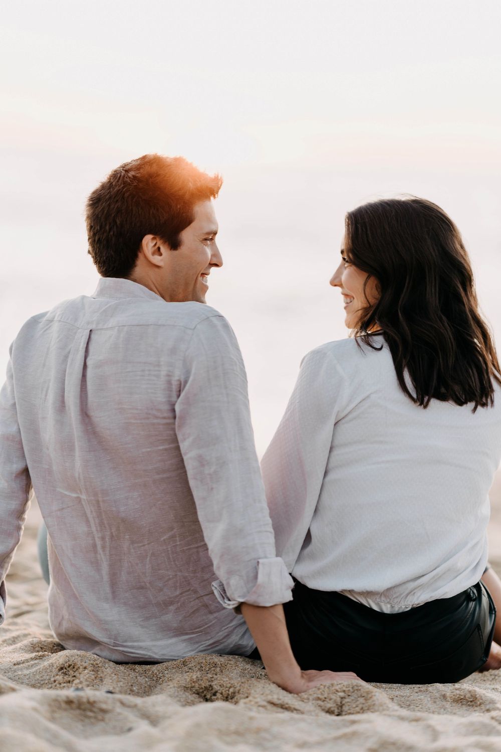 Couple in white shirts sitting together on beach sand at sunset sharing an intimate moment.