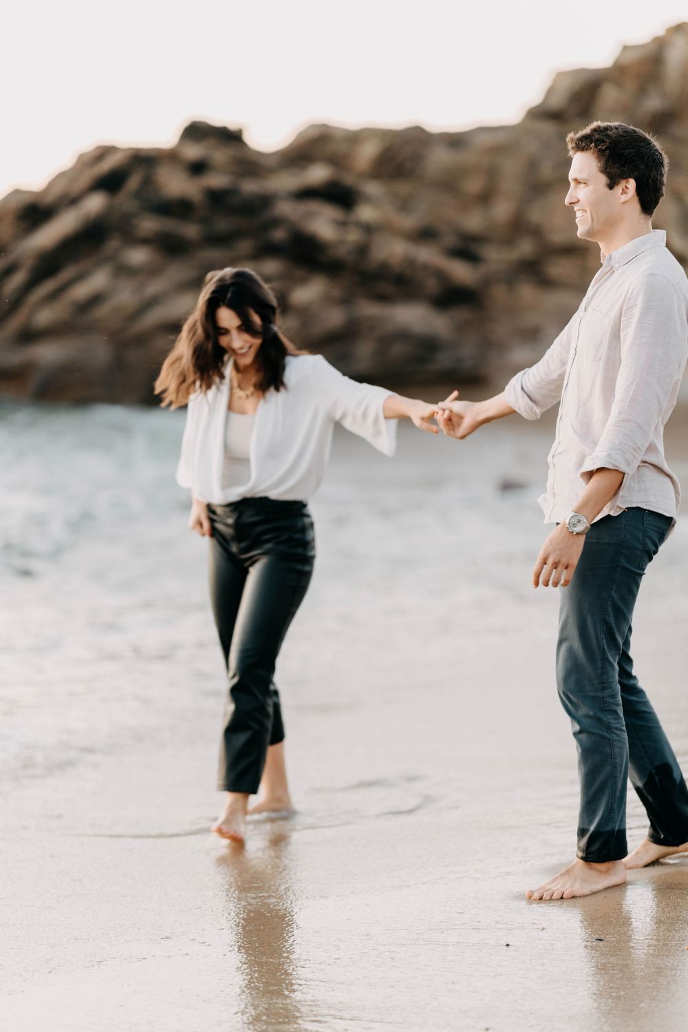 A couple walks playfully along the beach shoreline in white and dark casual attire during sunset.