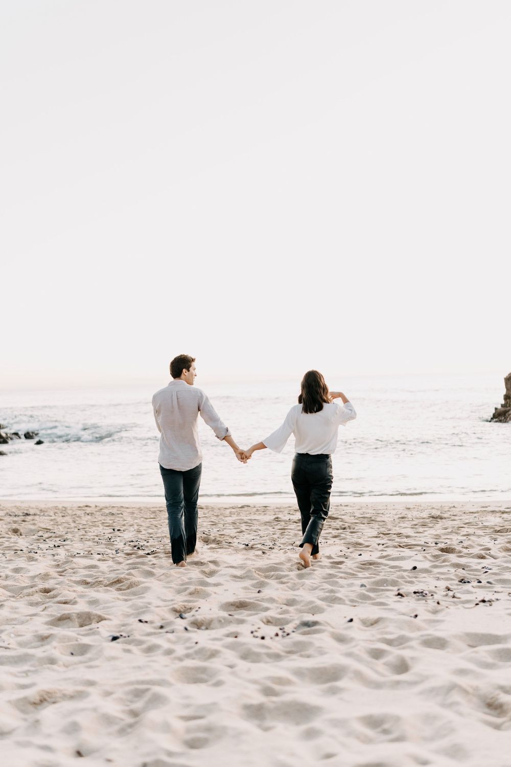 A couple runs playfully together across a white sandy beach on a bright sunny day.
