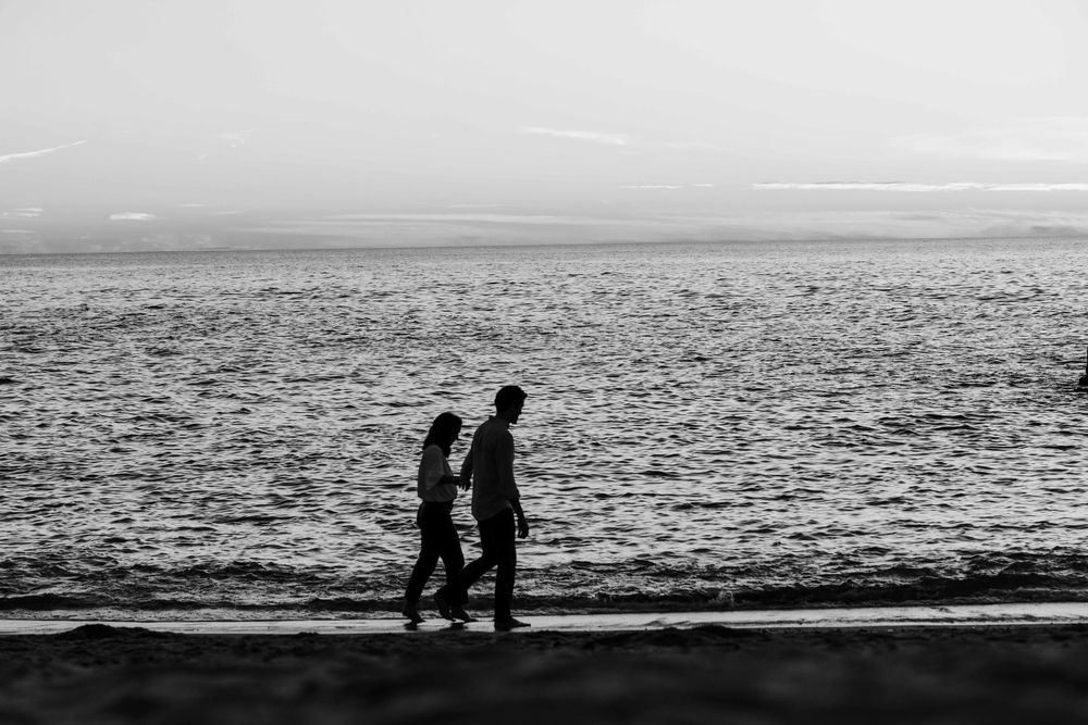 Two silhouetted figures walk along a beach shoreline at sunset with the ocean stretching to the horizon.