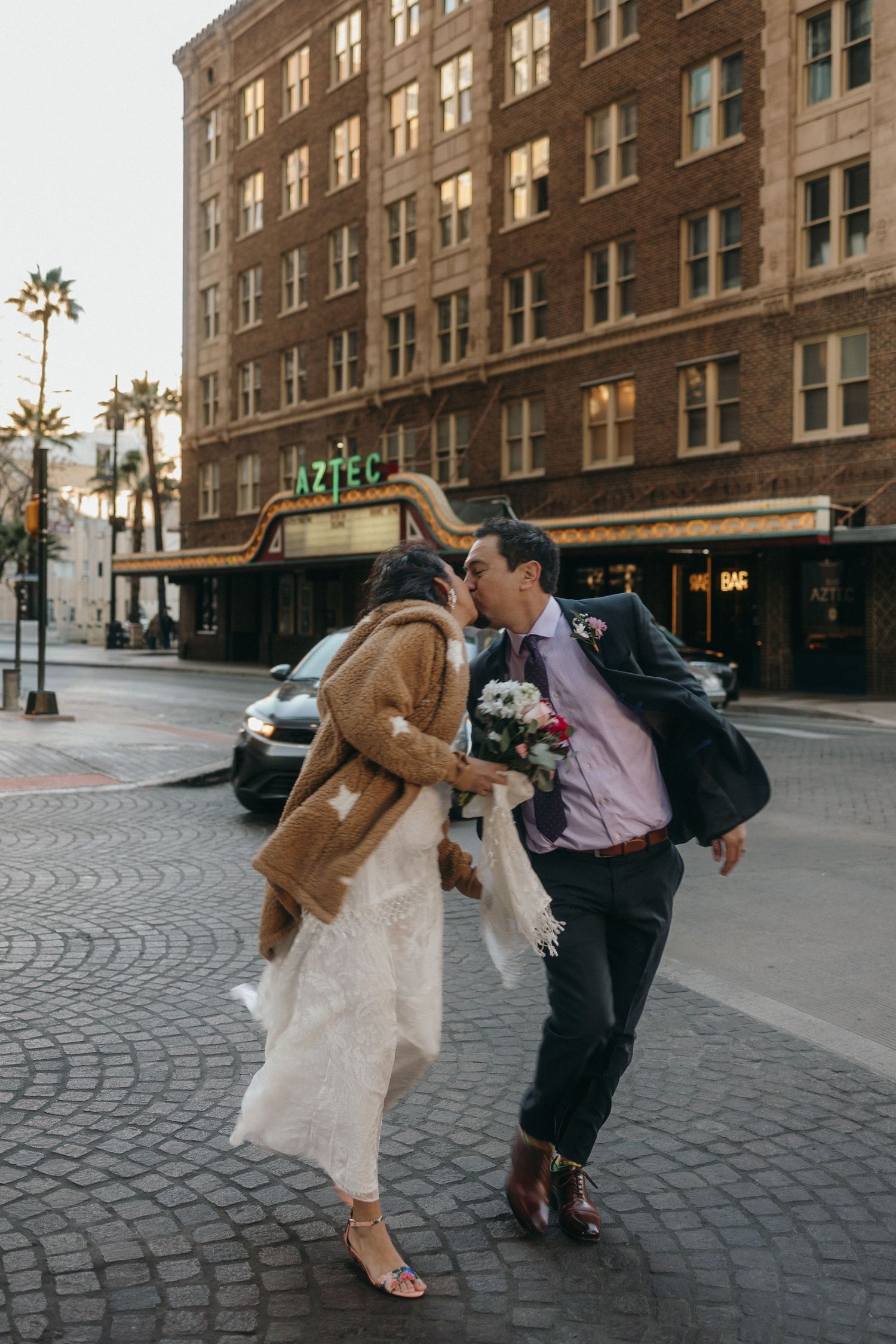 A romantic couple in formal attire dances playfully on a cobblestone street in front of a historic brick building.