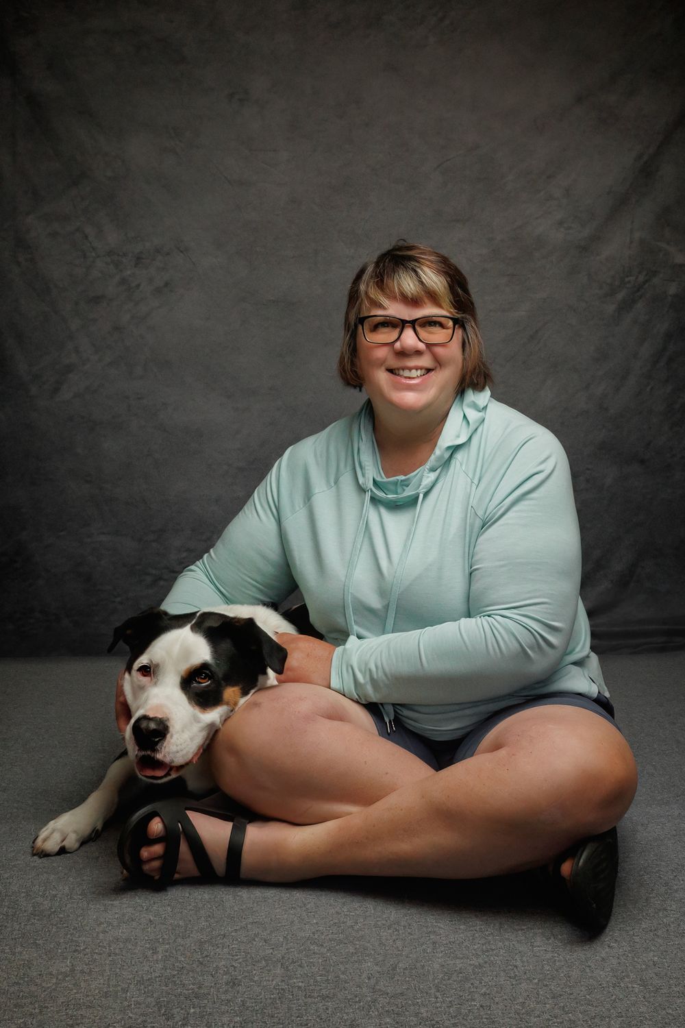 Smiling woman with glasses in mint top sits cross-legged on floor hugging a black and white dog against gray backdrop.