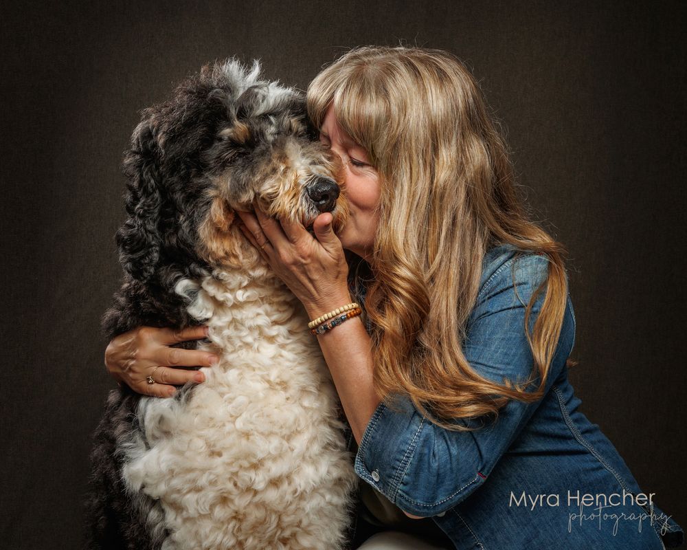 Woman in denim shirt kissing a fluffy black and white dog in a professional studio portrait by Myra Hencher Photography.