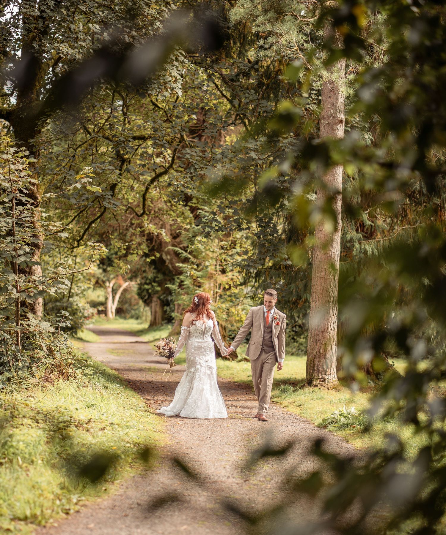 A couple walks together on a winding dirt path through a sunlit forest with green foliage overhead.