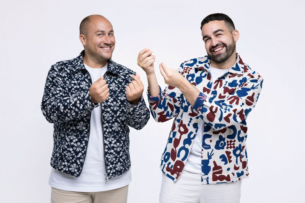 Two people wearing patterned jackets posing playfully together against white background.