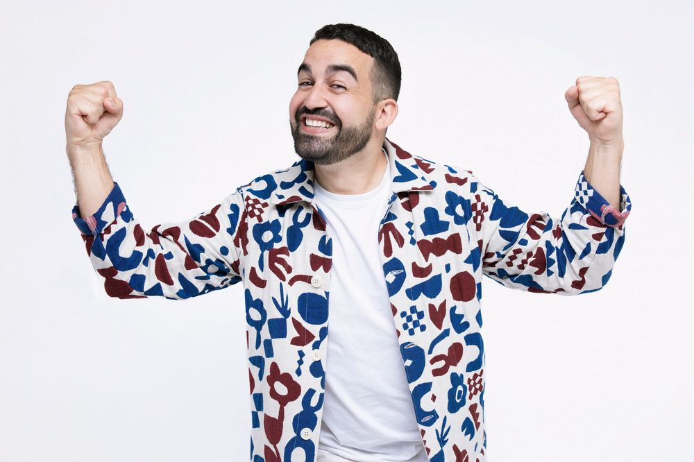 Man in colorful patterned shirt celebrating with raised arms against white background.