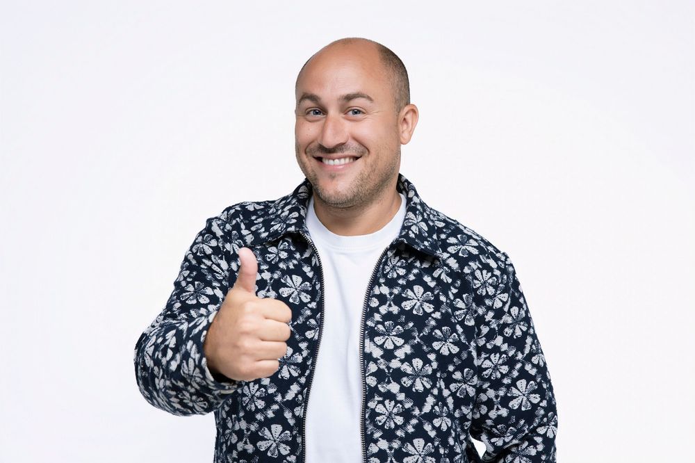 Person wearing floral print jacket and white shirt poses with thumbs up against white backdrop.
