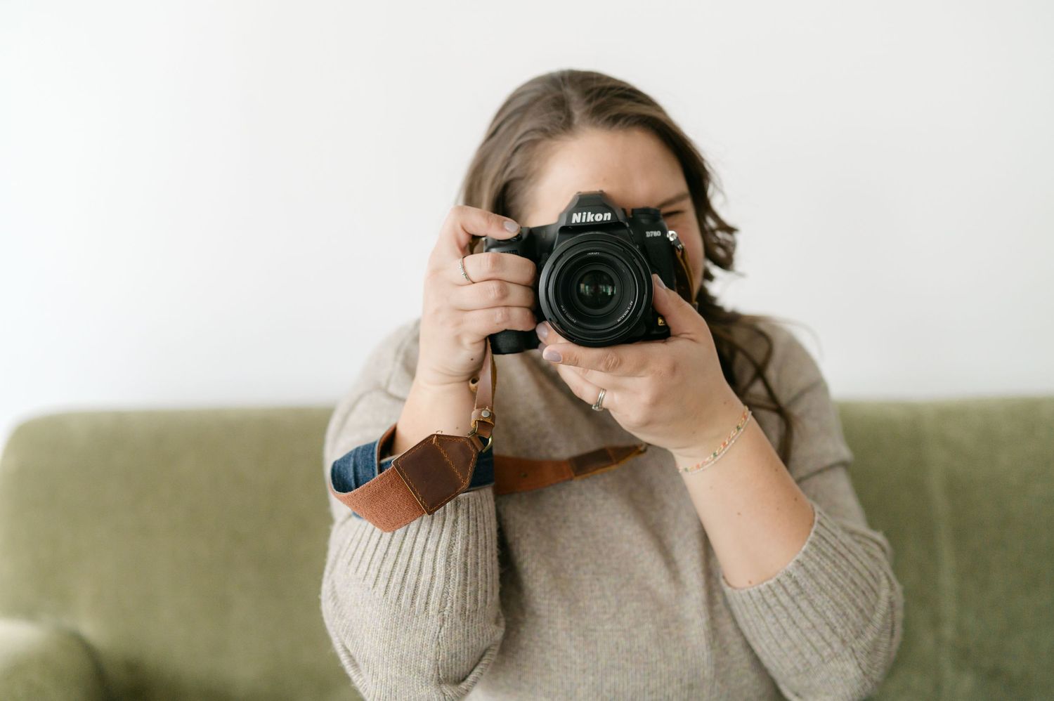 A person holds up a black DSLR camera in front of their face while sitting on a beige couch.