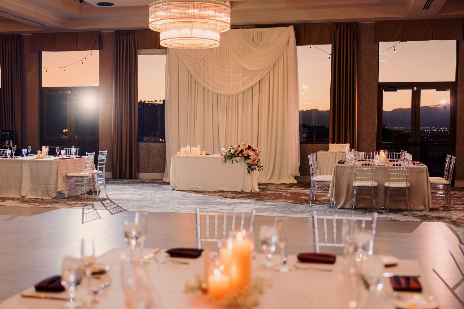 Elegant wedding reception venue with draped backdrop, chandelier, chiavari chairs, floral centerpiece, and mountain views at dusk.