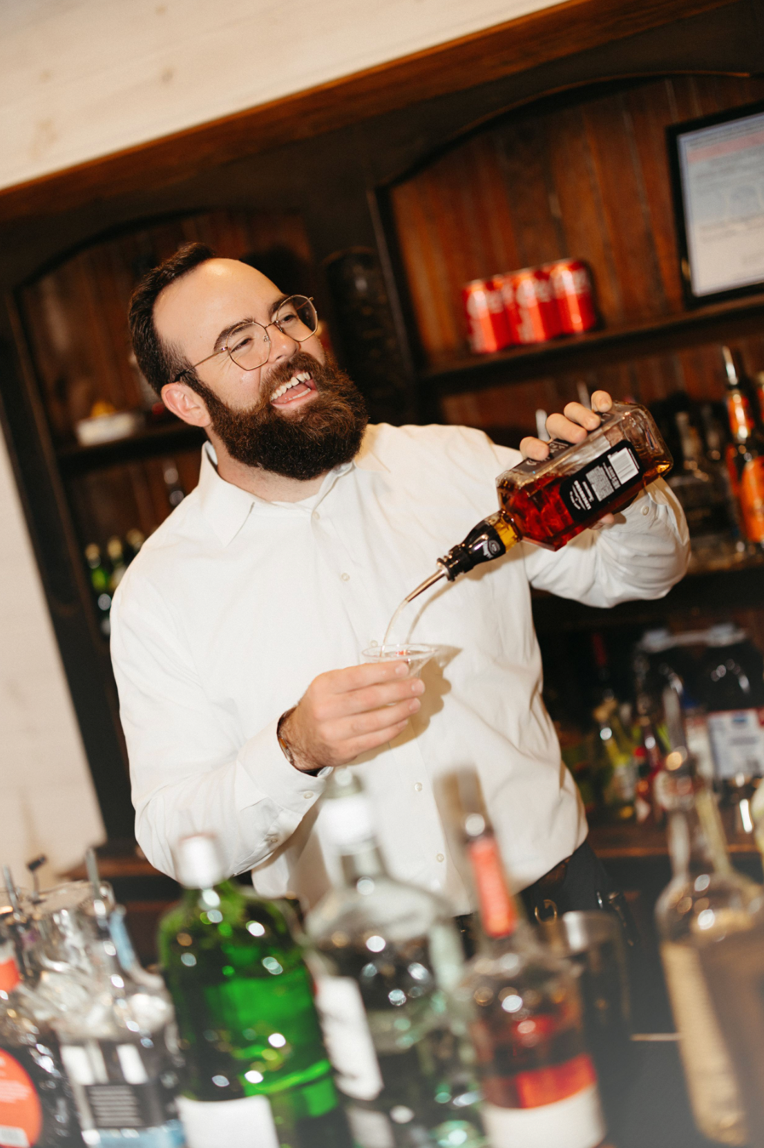Bartender in white shirt shaking cocktails with dramatic motion at a dimly lit bar counter.