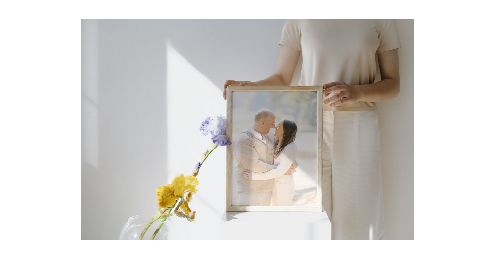 A sunlit interior scene showing hands holding a framed photograph with a yellow flower in a vase.