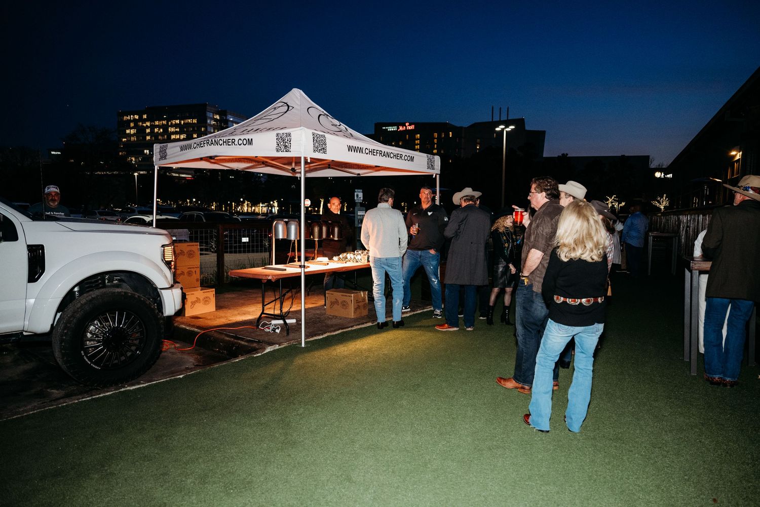 A nighttime scene showing a row of cars and vendor tents at an outdoor event under dark blue skies.