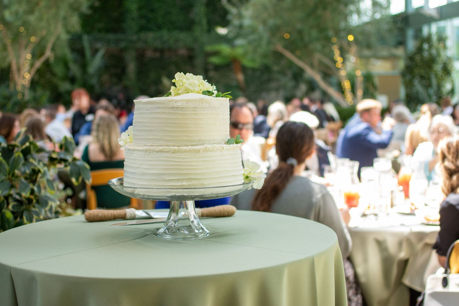 A white frosted wedding cake displayed at an outdoor garden reception with guests seated at tables in the background.