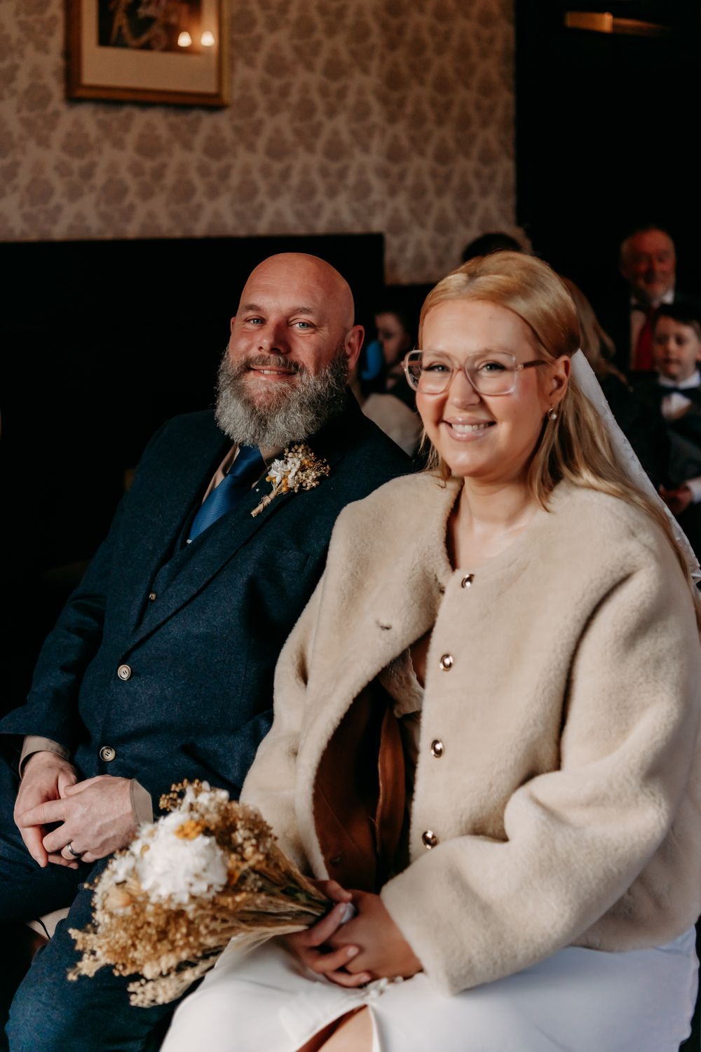 Wedding guests seated together in formal attire share warm smiles and laughter during an indoor celebration.