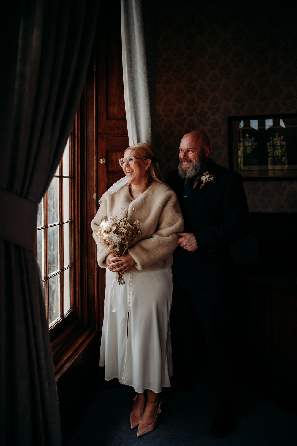 A couple in a dimly lit room wearing white, standing near curtained windows in a romantic indoor setting.