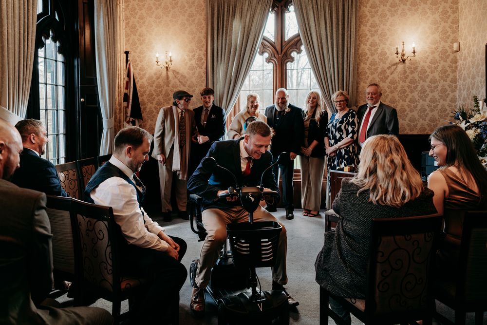 Wedding guests gather in an elegant ballroom with ornate wallpaper and chandeliers during an evening reception.