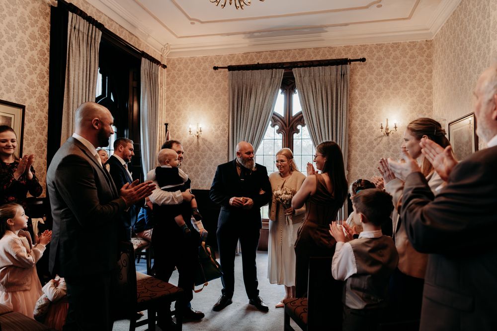 A wedding party dances and celebrates in an elegant ballroom with ornate crown molding and chandeliers.