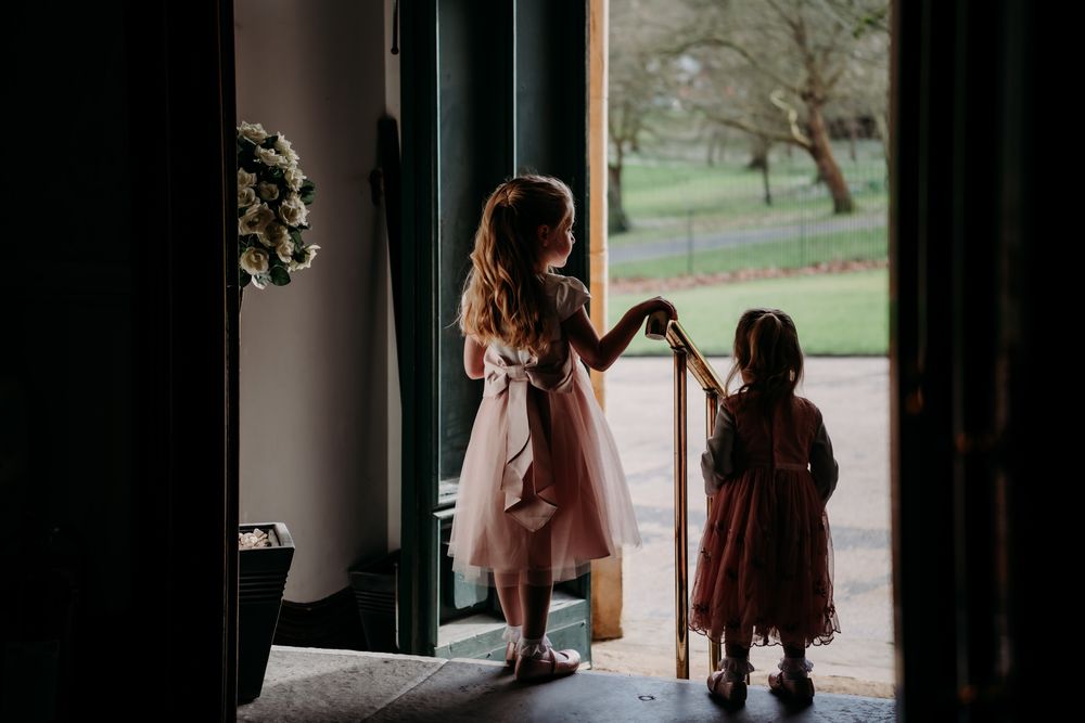 Two figures in dresses stand in a doorway silhouetted against natural light streaming in from outside.