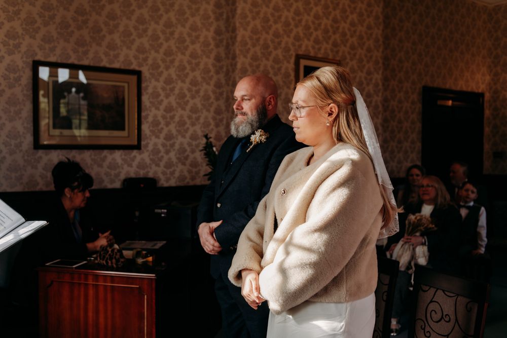 A couple stands together during a winter wedding ceremony in a room with patterned wallpaper.