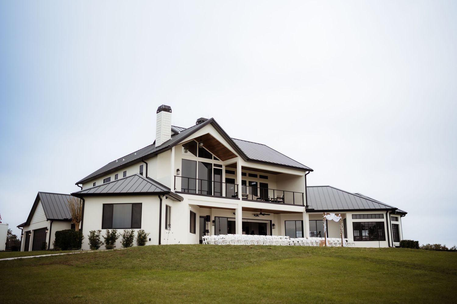 Modern white ranch-style home with large windows and wraparound porch sits atop a grassy hillside.