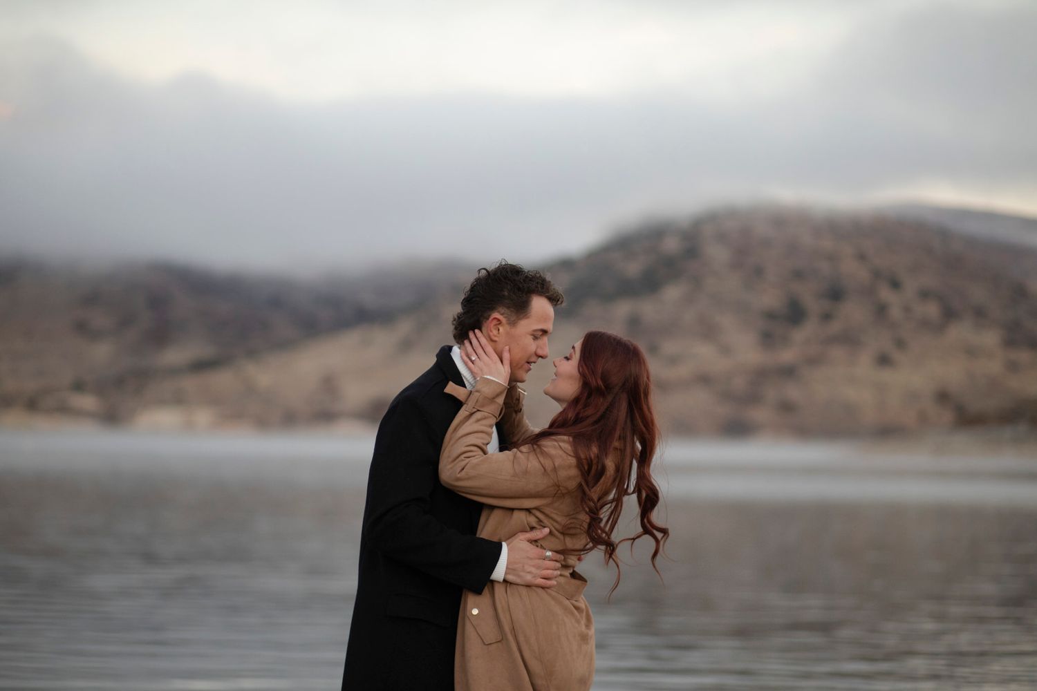 Couple stands in front of foggy mountains, facing each other, staring into each other's eyes.