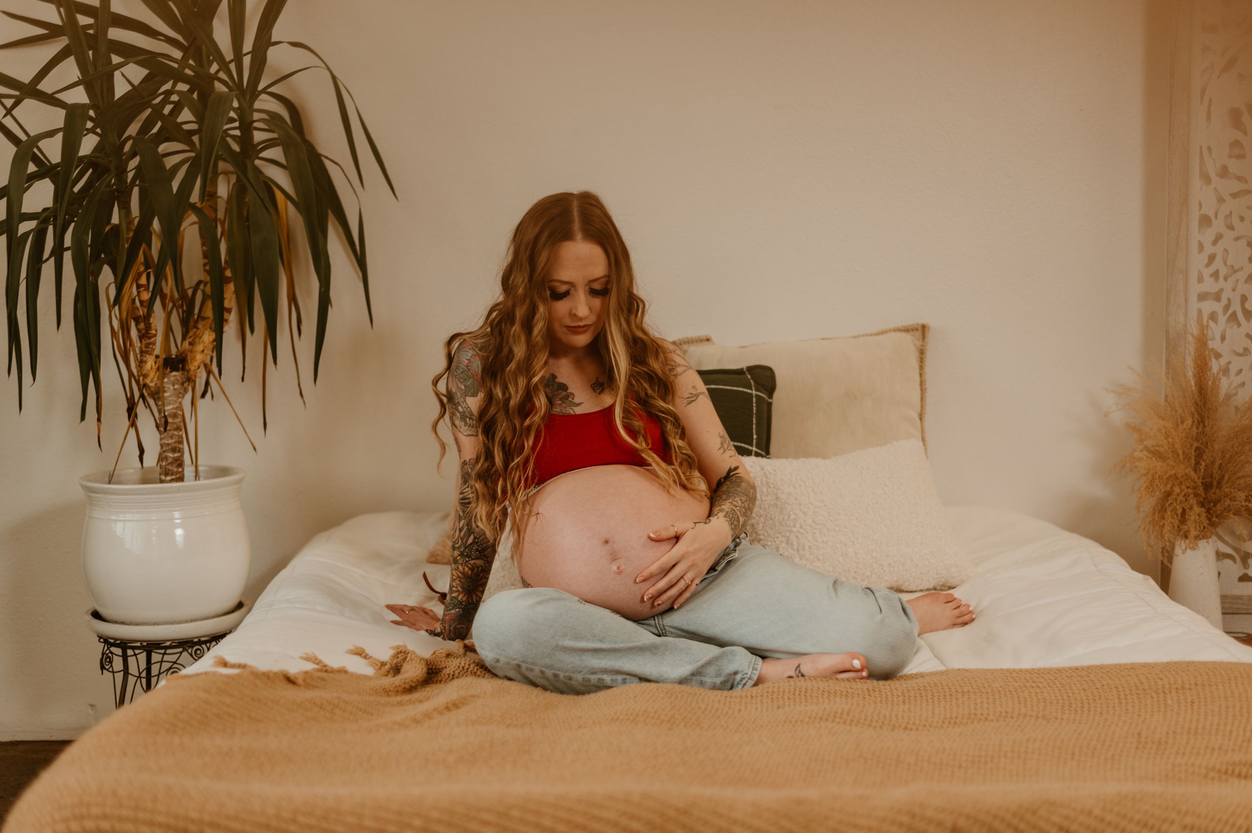 Expectant woman in red top relaxes on bed with decorative plant in warm-toned maternity photo.