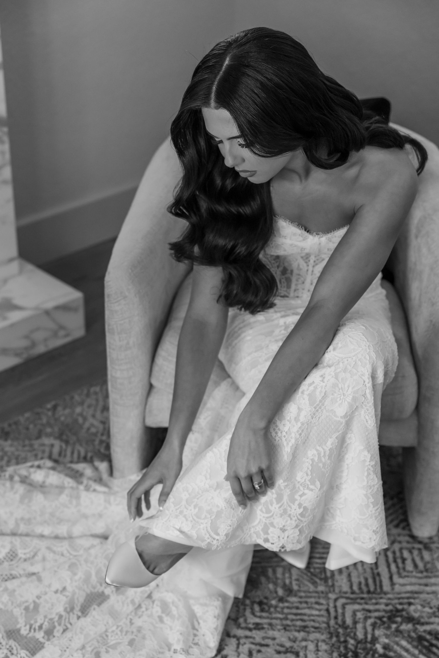 Black and white photo of a bride in a lace wedding dress holding a white rose bouquet next to a classic car.