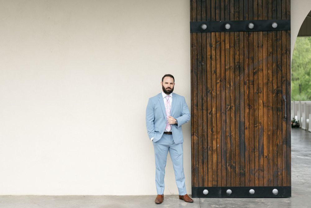 A groom in a powder blue suit poses confidently against a rustic wooden door.