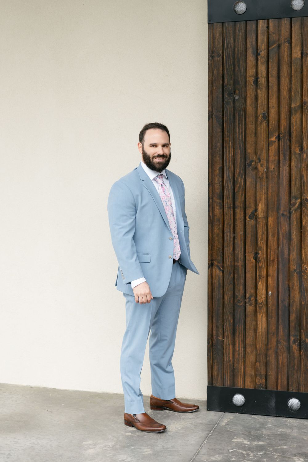 A groom in a light blue suit and brown shoes stands against a wooden accent wall.