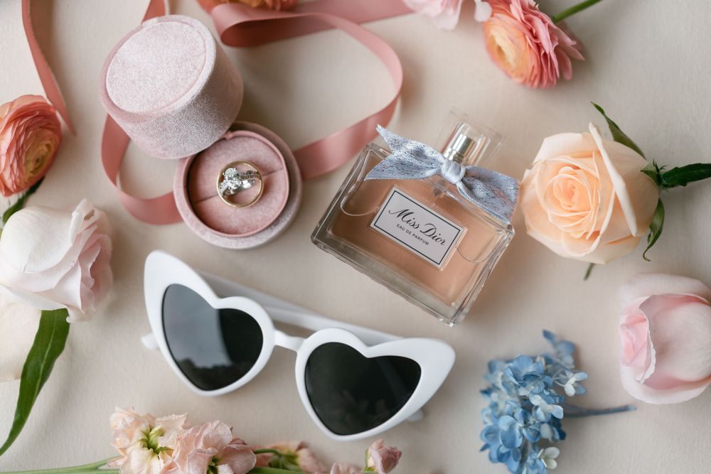 Wedding accessories flatlay with heart sunglasses, perfume, roses and ring box in pink tones.