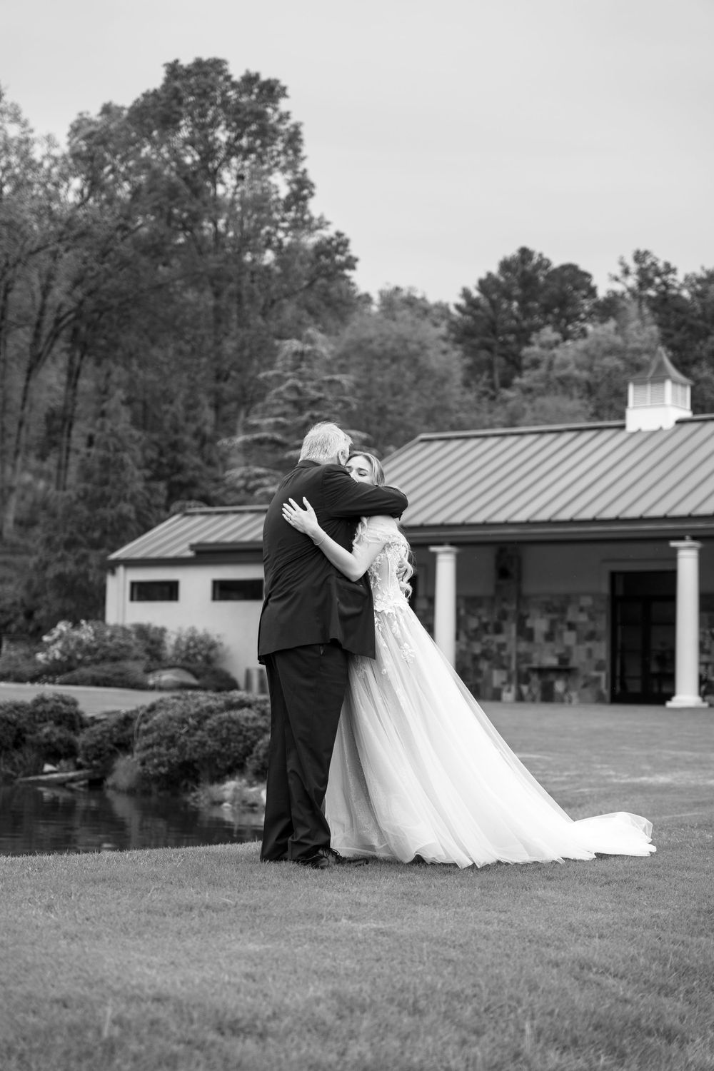 Black and white wedding photo with couple embracing on lawn with building in background.