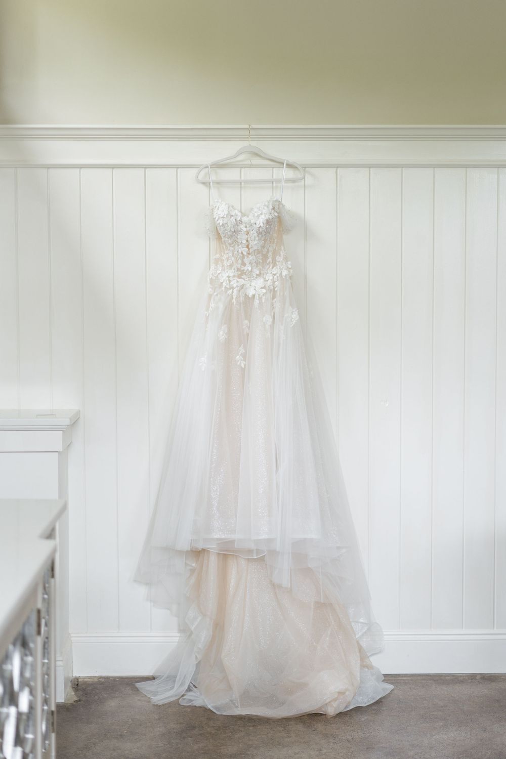 White lace wedding dress with tulle skirt hanging on display against white wainscoting.