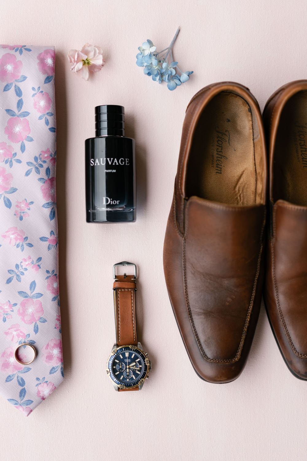 Groom's accessories including floral tie, brown leather shoes, cologne and watch on pink background.