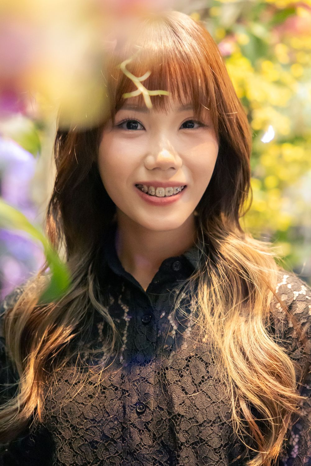 A person with long hair smiles among flowers wearing black lace top.