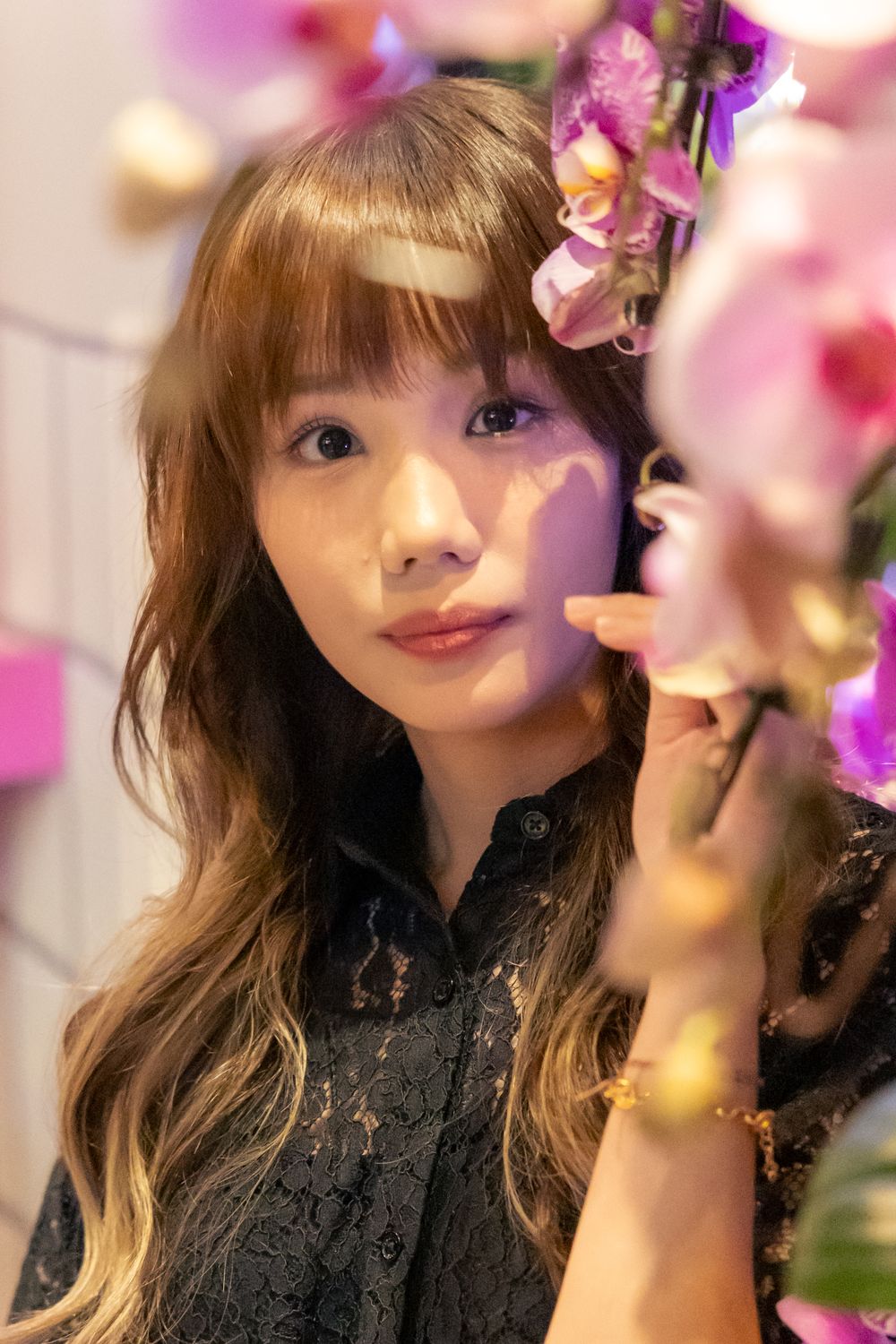 A person poses among purple orchids wearing black lace top with soft lighting.